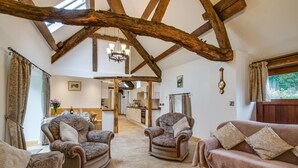 Un vaste espace de vie doté de hauts plafonds et de poutres apparentes, comprenant deux fauteuils à motifs et un canapé, un lustre, et offrant une vue sur la cuisine et la salle à manger.