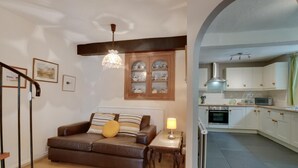 Una sala de estar con un sofá marrón, una mesita con una lámpara y una escalera. La cocina tiene armarios blancos y un salpicadero de azulejos.