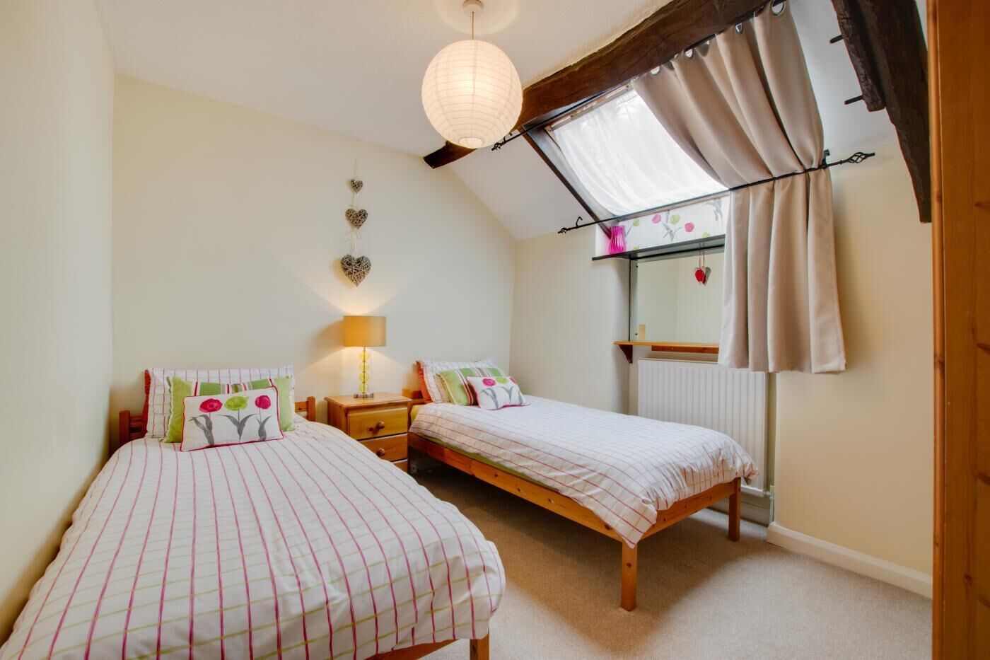 Un dormitorio con dos camas individuales, una mesita de noche con lámpara, una ventana con cortinas y una estantería en la pared con objetos decorativos.