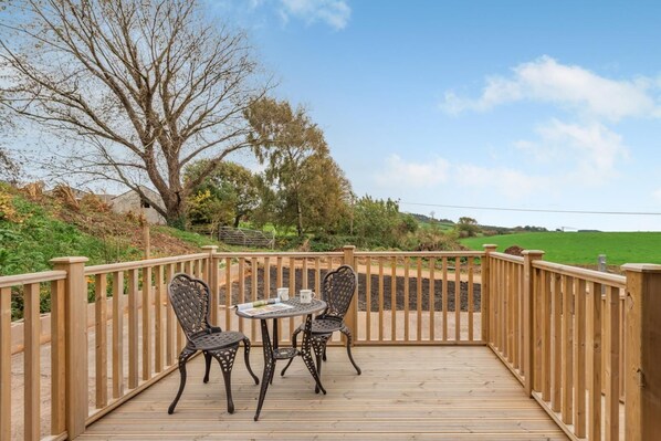 A wooden deck with a table and chairs, surrounded by a wooden railing, overlooking a green landscape.