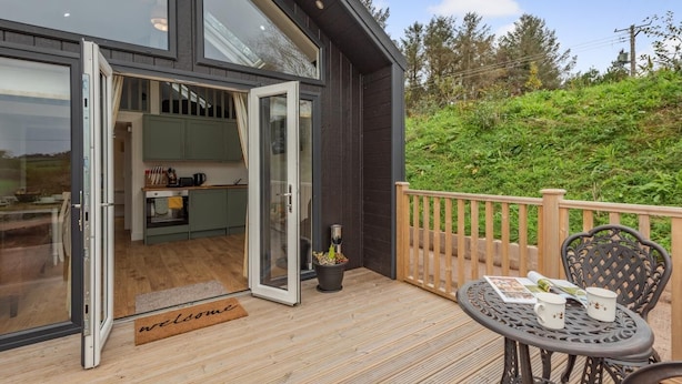 A modern house with a glass door leading to a kitchen and a wooden deck with a table and chairs. - 2 Bed in Mouswald (oc-b33444) (Mouswald)