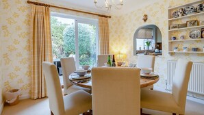 Zona pranzo con tavolo rotondo, sedie e lampadario. Si vede una finestra con tende, uno scaffale con oggetti decorativi e vista sul verde circostante.