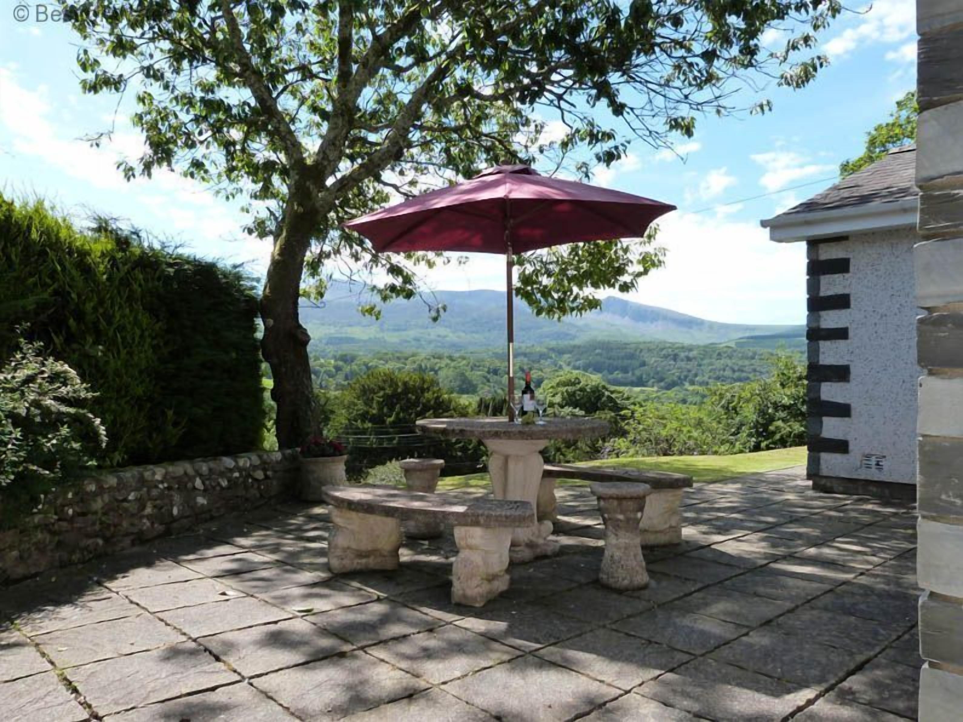 Un patio de piedra con mesa y sillas, una sombrilla roja y vistas a un paisaje montañoso.