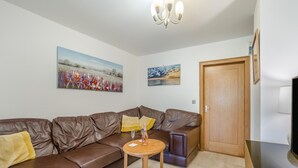 Uma sala de estar com um sofá de couro marrom, uma pequena mesa de madeira com uma garrafa e um copo, e duas pinturas na parede.