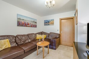 A living room with a brown leather sofa, a small wooden table with a bottle and glass, and two paintings on the wall. - 2 Bed in Cardiff (oc-45378) (Peterston-super-Ely)