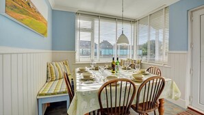 A dining area with a table set for four, white paneled walls, a bench with striped cushions, and a view through large windows. - 4 Bed in Old Hunstanton (oc-1809) (Hunstanton)