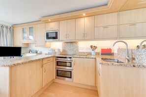 A modern kitchen with wooden cabinets, a microwave, two ovens, granite countertops, and a stainless steel sink. - 1 Bed in Saundersfoot (oc-pw9247) (Saundersfoot)