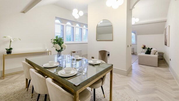 Un coin repas avec une table en verre, des chaises blanches et un vase de fleurs. Un grand miroir orne le mur, et une fenêtre est équipée de stores.