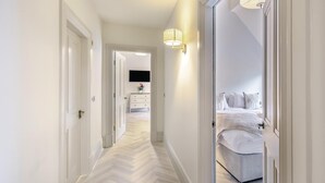 Un couloir au sol en parquet à chevrons menant à une chambre à coucher équipée d’un lit et d’une table de chevet.
