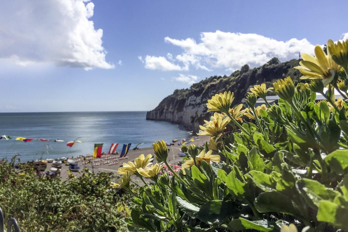 A coastal beach scene with a rocky cliff, clear sky, and colorful flags.