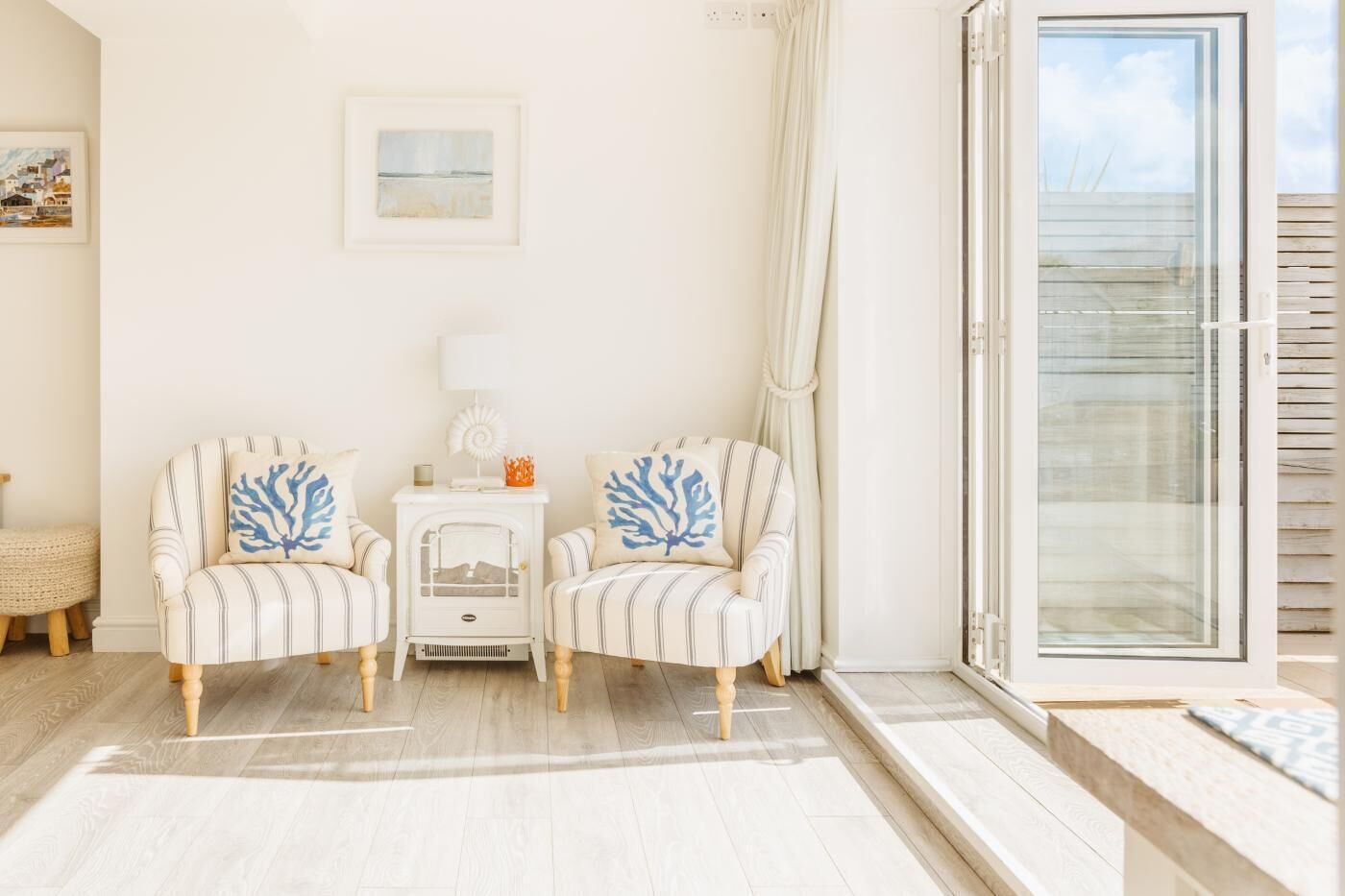 Une pièce avec deux fauteuils à rayures, une petite table blanche, une lampe blanche et une porte en verre donnant sur l’extérieur.