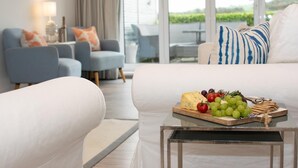 A modern living room with a white sofa, a coffee table with a cheese platter, and a view of a garden through large windows. - 4 Bed in Little Petherick (oc-urchpe) (Little Petherick)