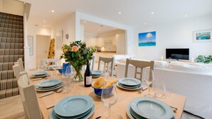 A modern dining area with a table set for four, a vase of flowers, and a kitchen in the background. - 4 Bed in Little Petherick (oc-urchpe) (Little Petherick)