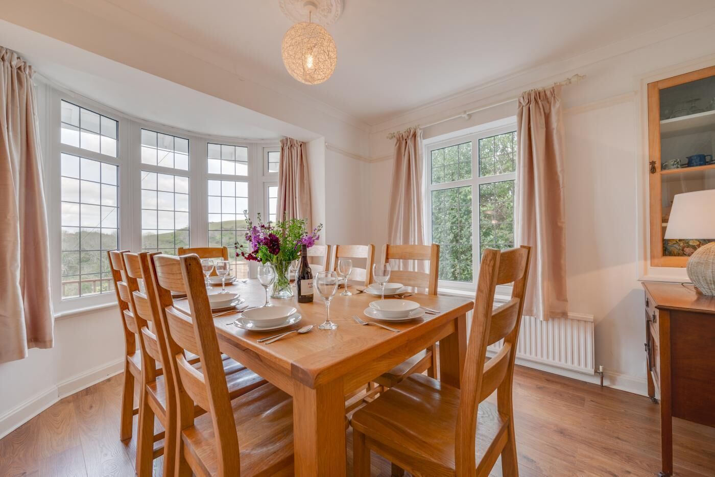 Une salle à manger avec une table en bois dressée pour six personnes, de grandes fenêtres et un lustre.