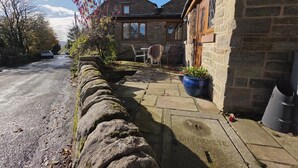 A stone house with a paved driveway, a stone wall, and a garden with a blue pot.
