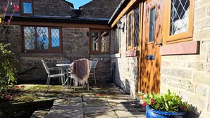 A stone building with a wooden door and windows. There is a table and chairs outside, and a blue planter with flowers.