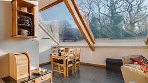 A dining area with a wooden table and chairs, a wooden cabinet with decorative items, and a large window with a view of trees. - 2 Bed in Dolgellau (oc-bow282) (Blaenau Ffestiniog)
