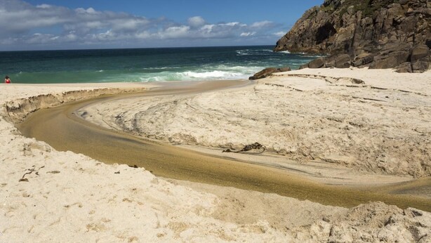 Ein sandiger Strand mit einem gewundenen Pfad zum Meer, felsigen Klippen und einem wolkenlosen Himmel.