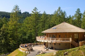 Eine Holzhütte mit einem Balkon und einer runden Sitzecke, umgeben von bewaldeter Landschaft.