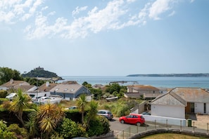 A coastal residential area with houses, a red car, and a hill with a castle-like structure in the distance. - 4 Bed in Marazion (oc-gwynsa) (Marazion)