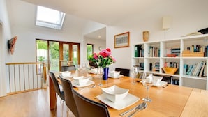 A dining room with a long wooden table set for six, surrounded by chairs, with a bookshelf and a window with a view of greenery. - 4 Bed in Marazion (oc-gwynsa) (Marazion)