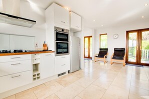 A modern kitchen with white cabinetry, a built-in oven, and a stove. There are two chairs in a sunlit area with a view of greenery outside. - 4 Bed in Marazion (oc-gwynsa) (Marazion)