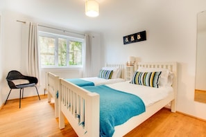 A bedroom with a white bed, two pillows, a blue blanket, a black chair, a window with curtains, and a wall-mounted light fixture. - 4 Bed in Marazion (oc-gwynsa) (Marazion)
