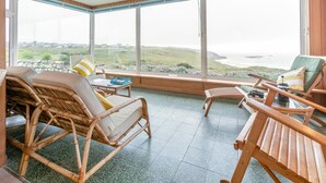 Ein Zimmer mit großen Fenstern, Korbmöbeln, einem kleinen Tisch und Meerblick.