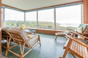 Une pièce dotée de grandes fenêtres, de meubles en osier, d’une petite table et offrant une vue sur la mer.