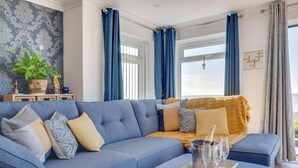A modern living room with a blue sofa, a wooden coffee table with a tray of glasses and a bottle, and a view of the sea through large windows.