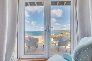 Une pièce dotée d’une porte-fenêtre coulissante donnant sur une terrasse extérieure avec deux chaises et une vue sur la mer et les collines.