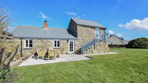 A stone house with a staircase leading to a second floor, a grassy yard, and a small outdoor seating area with wooden chairs. - 3 Bed in Sennen (oc-thbyre) (Sennen)