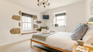 A bedroom with a bed, two windows with stone walls, a chandelier, a bedside table with a book, and a nightstand with a lamp. - 3 Bed in Penzance (oc-songpz) (Penzance)