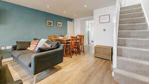 A living room with a grey sofa, wooden dining table, and chairs. A staircase is visible on the right. - 3 Bed in Penzance (oc-songpz) (Penzance)