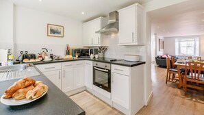A modern kitchen with white cabinets, a built-in oven, and a dining area with wooden chairs and a table. - 3 Bed in Penzance (oc-songpz) (Penzance)