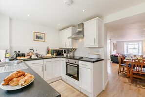 A modern kitchen with white cabinets, a built-in oven, and a dining area with wooden chairs and a table. - 3 Bed in Penzance (oc-songpz) (Penzance)