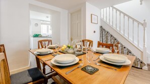 A dining area with a wooden table set for four, white walls, a staircase, and a kitchen visible in the background. - 3 Bed in Penzance (oc-songpz) (Penzance)