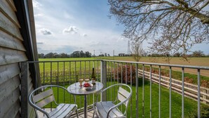 A balcony with two metal chairs and a small round table, overlooking a green field and a tree. - 1 Bed in Booton (oc-2266) (Cawston)
