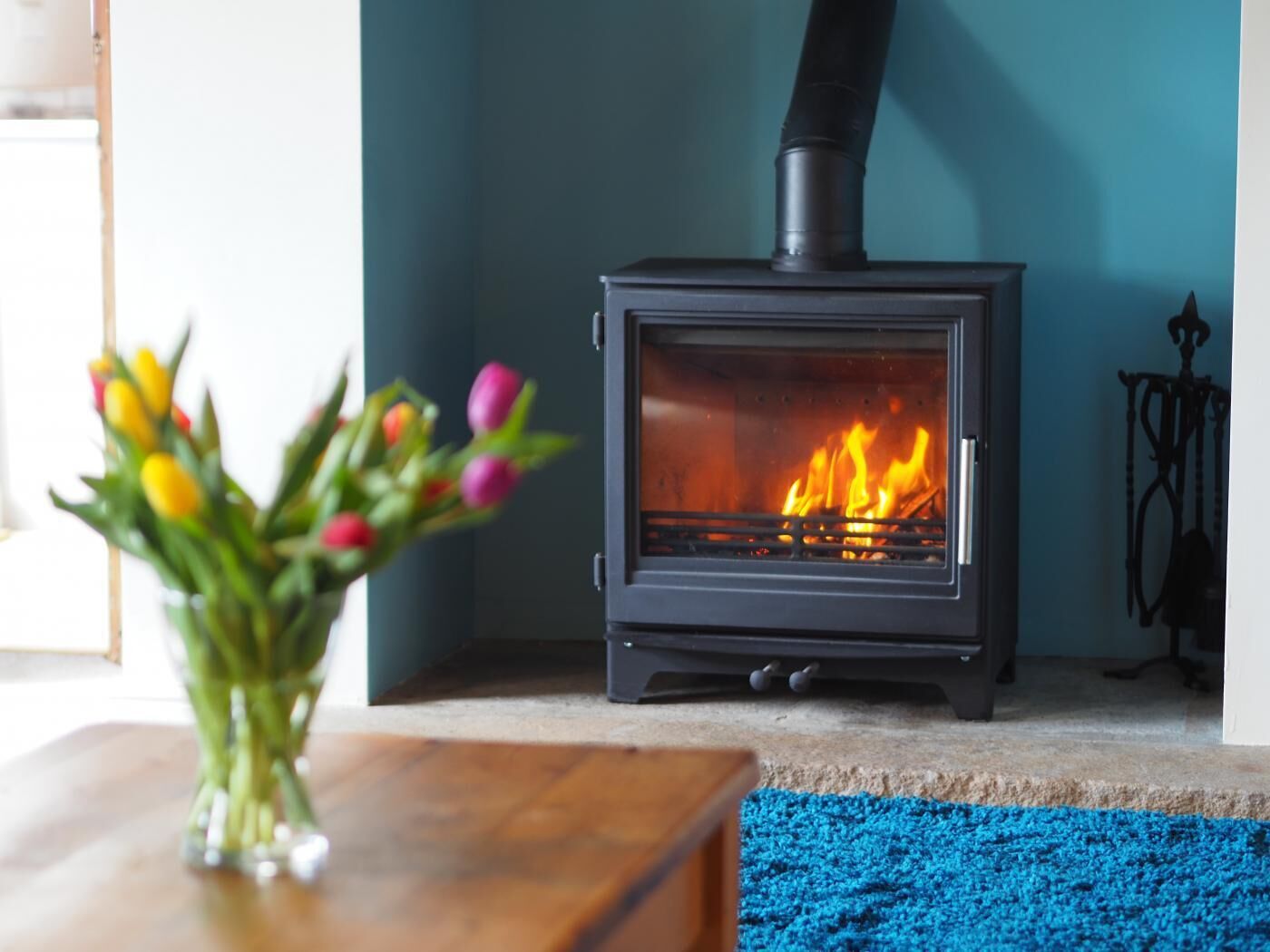 Ein Kaminofen mit sichtbarem Feuer, eine Vase mit Tulpen und ein blauer Teppich auf einem Holztisch.