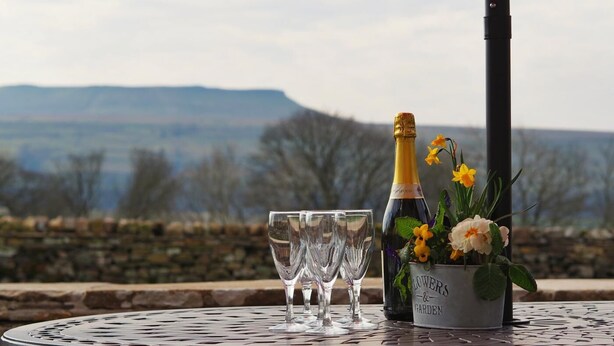 A table with a bottle of champagne, two glasses, and a flower arrangement on a metal table with a scenic view in the background. - 3 Bed in Askrigg (oc-ds1049) (Askrigg)