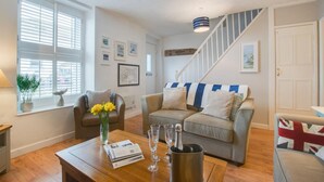 A living room with a sofa, a coffee table with magazines and glasses, a vase with flowers, and a staircase with a striped cushion. - 2 Bed in Mousehole (oc-nigelh) (Mousehole)