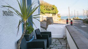 A seaside patio with wicker furniture, a white wall with the name "NIGEL", and a view of the sea. - 2 Bed in Mousehole (oc-nigelh) (Mousehole)
