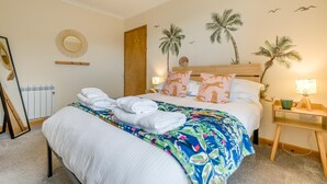 Una cama bien hecha con sábanas blancas y una colcha floral colorida, una mesita de noche de madera con una lámpara, y una pared decorada con motivos de palmeras.
