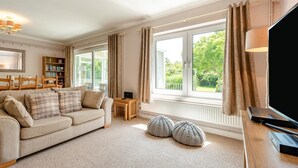 A living room with a sofa, a TV, a bookshelf, and large windows with curtains. - 2 Bed in Port Eynon (oc-hh2104) (Port Eynon)
