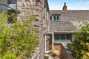 Une maison en pierre avec un petit porche, une fenêtre ornée de volets bleus et une chaise à l’extérieur.