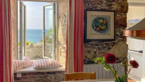 A room with a stone wall, a window with a view of the sea, a framed painting, and a table with a vase of flowers.