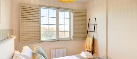 Ein Schlafzimmer mit einem Bett, Kissen, einem Fenster mit Jalousien, einem Heizkörper, einem Handtuchhalter und einer Hängeleuchte.