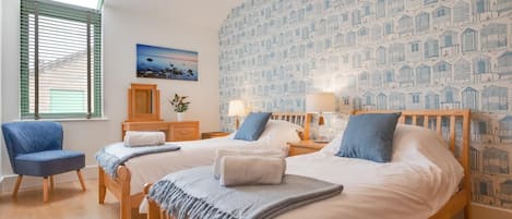 A bedroom with two beds, a chair, a dresser, a painting, and a patterned wallpaper.