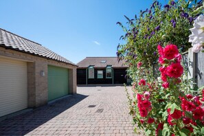 A courtyard with a paved walkway, a building with a tiled roof, and a flower bed with pink flowers. - 2 Bed in Holt (oc-2274) (Holt)