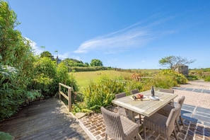 A wooden deck with wicker chairs and a table set for dining, overlooking a grassy field and a stone building. - 3 Bed in Mousehole (oc-kemyel) (Lamorna)